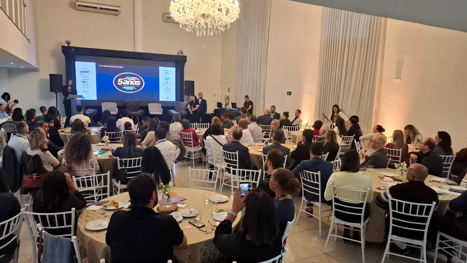 Vista ampla de um salão de eventos com mesas redondas cheias de pessoas, um palco com telão exibindo o logo ACIAS e patrocina