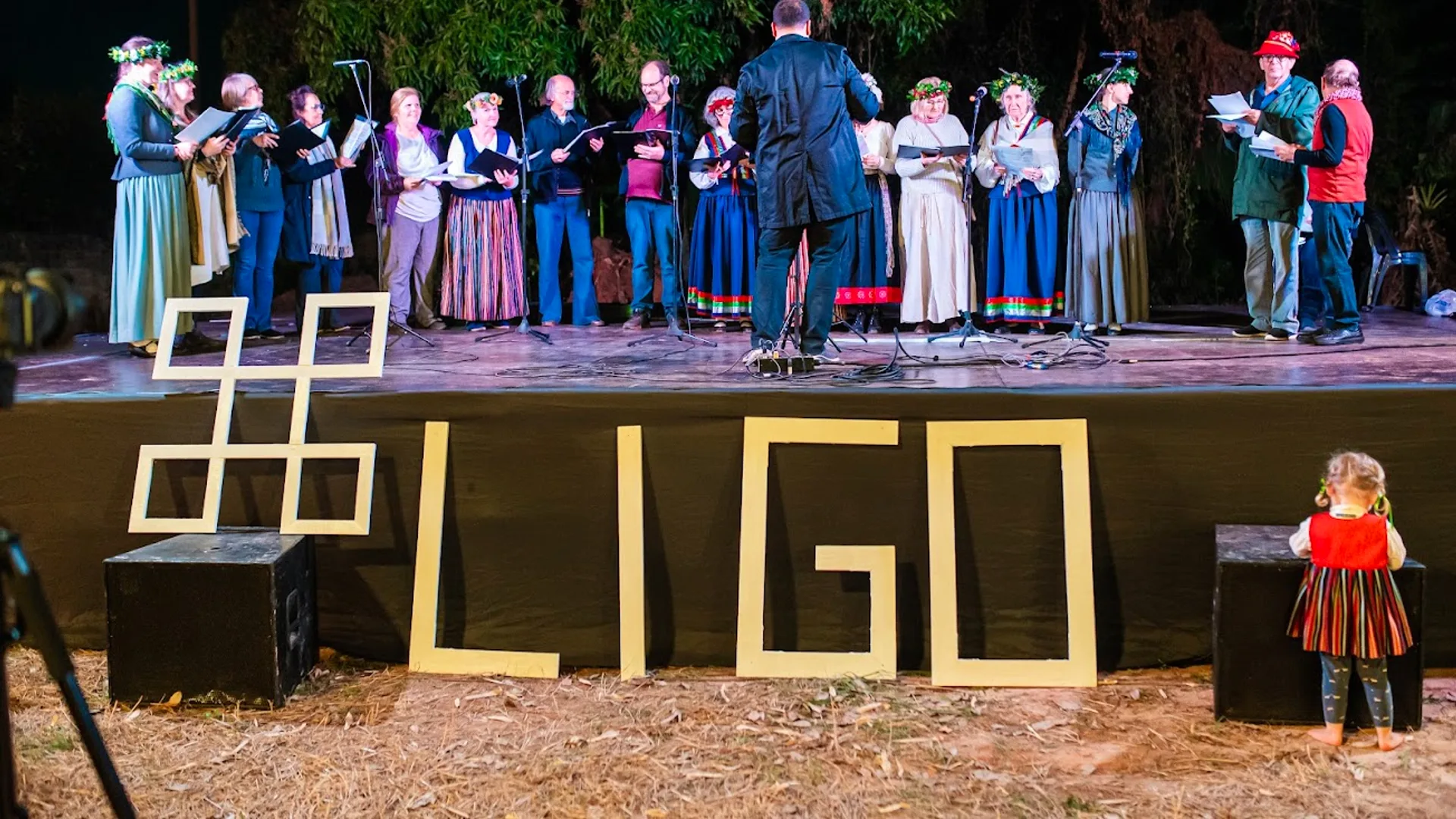 Coro se apresenta no palco com o nome 'LĪGO' em destaque, enquanto uma criança observa a celebração da cultura letã.
