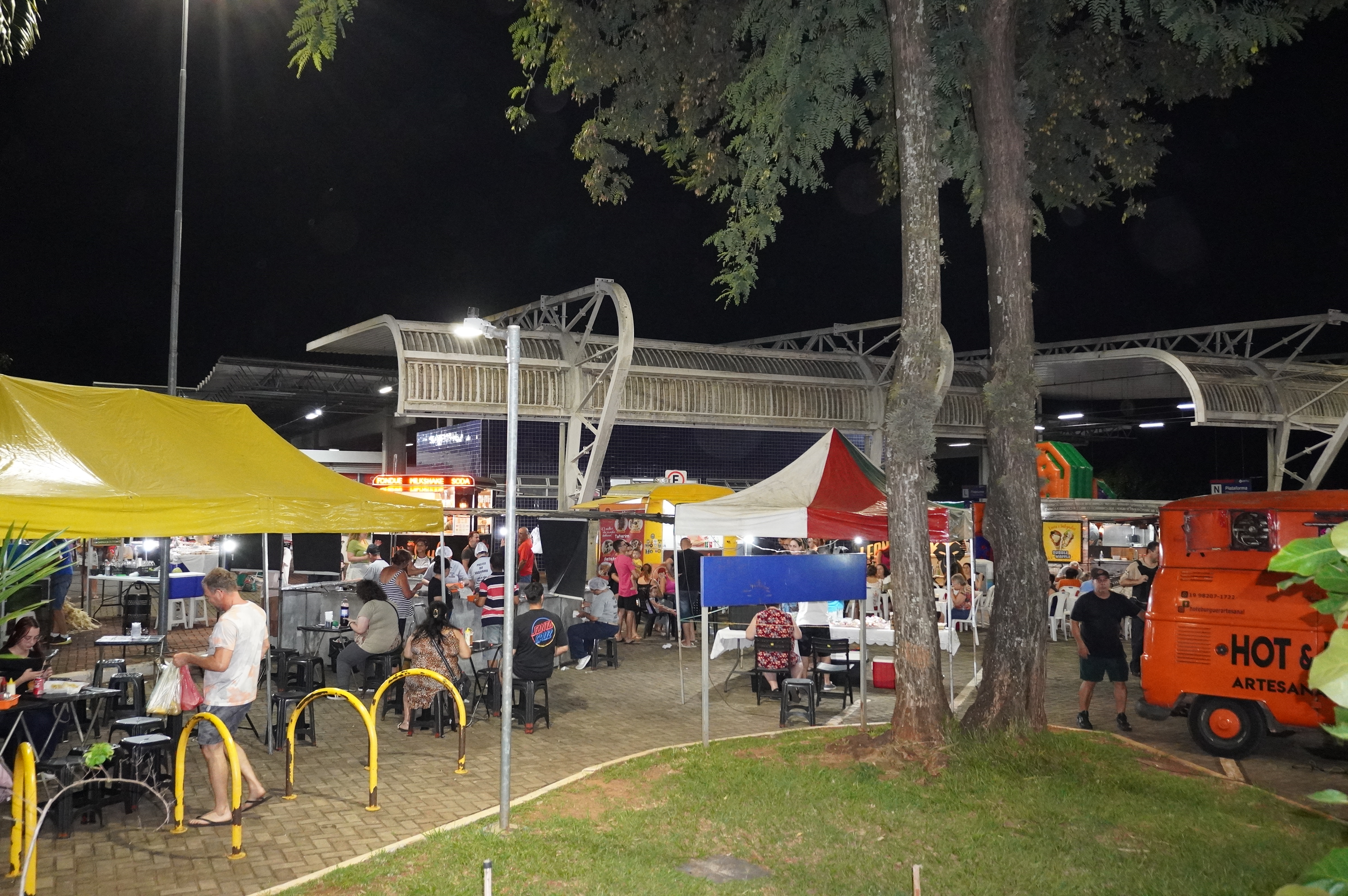 Feira noturna ao ar livre com barracas de comida e público circulando sob clima instável