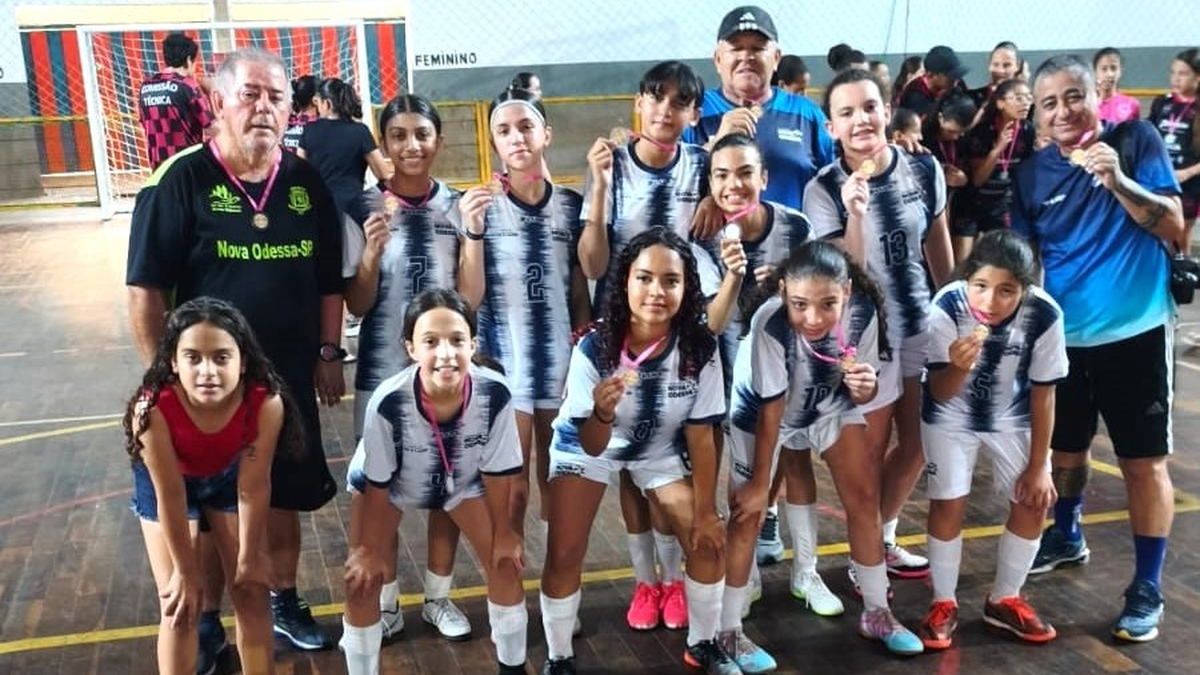Meninas da equipe Sub-13 de futsal de Nova Odessa reunidas com a comissão técnica exibindo troféu e medalhas.