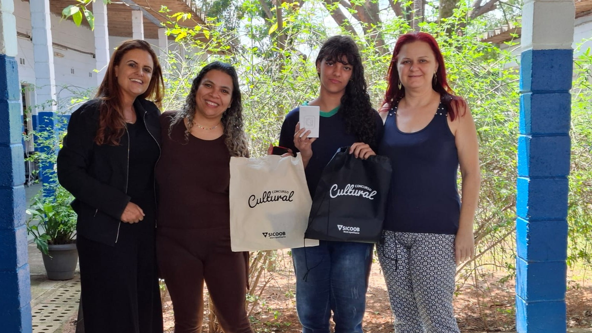 Estudante e educadoras seguram sacolas e prêmio do Concurso Cultural Sicoob em escola municipal de Sumaré